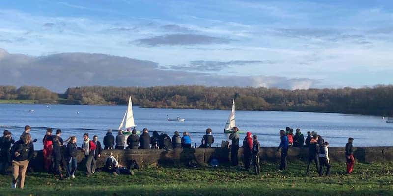 Two firefly dinghies in the distance on a reservoir with sailors watching from the shore in the foreground